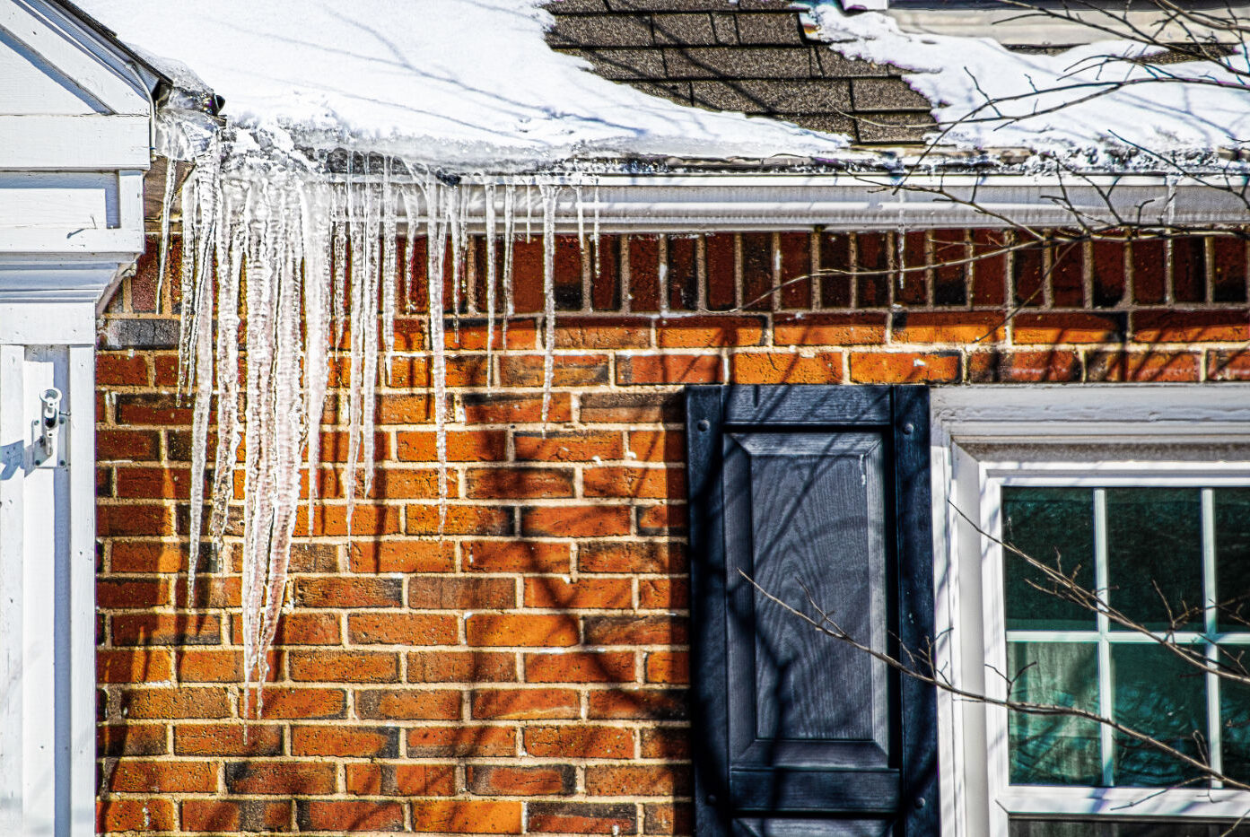 Large icicles and ice dams hanging from snow covered roof of brick house, indicating poor roof insulation.