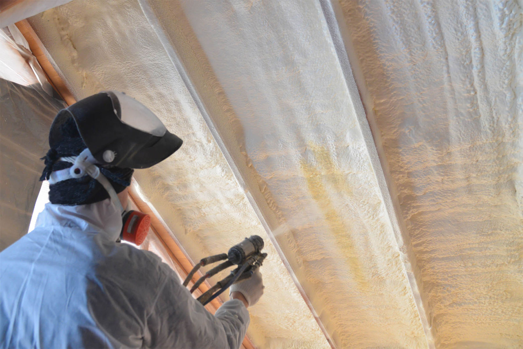 Worker in safety gear applying spray foam insulation to ceiling beams.