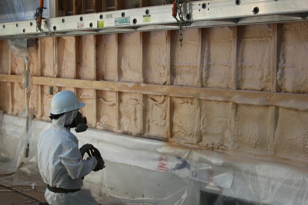 Worker in safety gear applying spray foam insulation to wall studs.