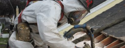 Worker in protective gear spraying foam insulation on roof boards.