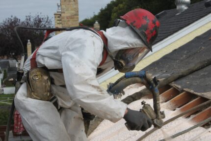 Worker in protective gear spraying foam insulation on roof boards.