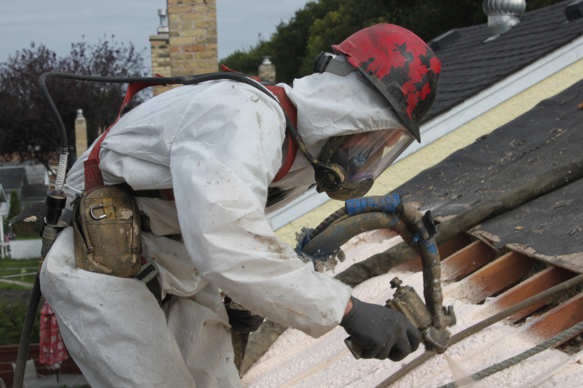 Worker in protective gear spraying foam insulation on roof boards.